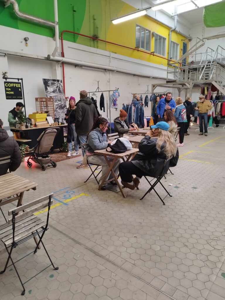 Bezoekers zitten bij de Kaai Markt aan tafeltjes met koffie en kleine hapjes, terwijl de markt met kraampjes en passerende bezoekers op de achtergrond zichtbaar is